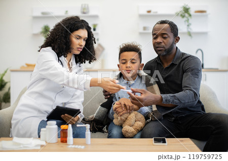 African female doctor assisting father and son with thermometer at home 119759725
