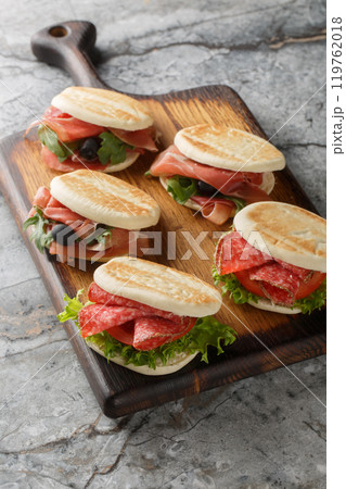 Popular street food in Emilia-Romagna, tigelle or crescentine sandwich with salami, ham and fresh vegetables close-up on wooden board. Vertical Popular street food in Emilia-Romagna, tigelle or crescentine sandwich with salami, ham and fresh vegetables close-up on wooden board. Vertical 119762018
