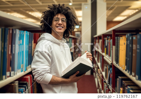Dark-haired young guy standing near the bookshelves and reading a book Dark-haired young guy standing near the bookshelves and reading a book 119762167