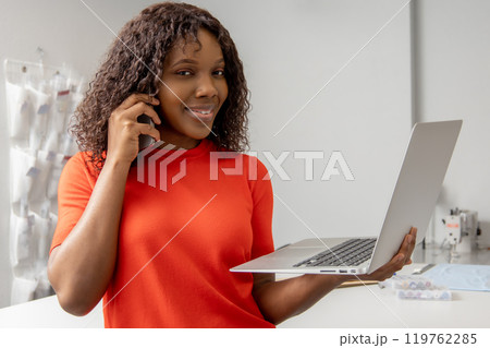 African american young seamstress with a laptop discussing something on the phone African american young seamstress with a laptop discussing something on the phone 119762285