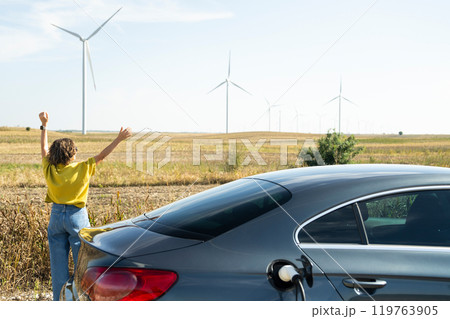 Woman with curve hairs wearing yellow shirt stands next to charging electric car. Wind turbines in the background Woman with curve hairs wearing yellow shirt stands next to charging electric car. Wind turbines in the background 119763905