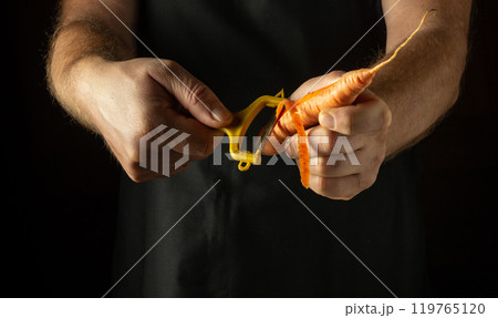 Vegetable peeler in the hand of a cook to clean carrots before preparing a vitamin salad. Low key concept of preparing vegetables for dinner Vegetable peeler in the hand of a cook to clean carrots before preparing a vitamin salad. Low key concept of preparing vegetables for dinner 119765120