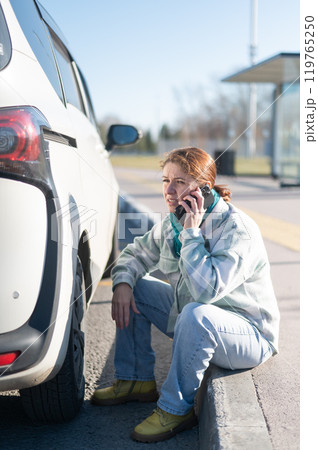 A Caucasian woman sits by a car wheel and calls for help on the phone. A Caucasian woman sits by a car wheel and calls for help on the phone. 119765250