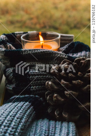 Orange scented wax candle in glass tumbler wrapped in a knitted scarf or sweater with a green natural background. Pine cone. Cozyness of slow living. Orange scented wax candle in glass tumbler wrapped in a knitted scarf or sweater with a green natural background. Pine cone. Cozyness of slow living. 119766182
