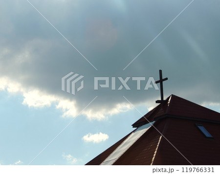 The tiled roof of a Protestant church with a cross on top against a blue sky with a large gray cloud in the background The tiled roof of a Protestant church with a cross on top against a blue sky with a large gray cloud in the background 119766331