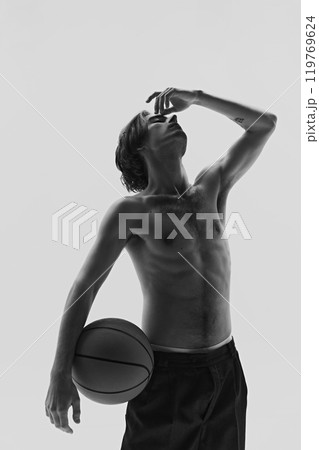 Young man holding basketball, hand raised to his face in thoughtful pose. Dynamic photo in monochrome filter. 119769624