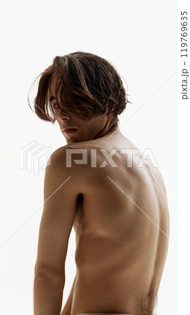 Portrait of young man posing shirtless and looking at camera over shoulder against white studio background. Male model revealing his bare back. Portrait of young man posing shirtless and looking at camera over shoulder against white studio background. Male model revealing his bare back. 119769635