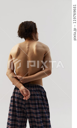 Back view of young man in plaid pants with hands behind back against white studio background. Shows natural physique, relaxation, and wellness. 119769681