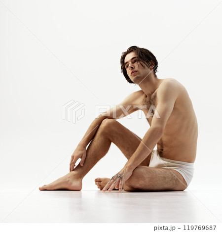 Young man seated casually, legs crossed, showcasing relaxed style and masculinity against white studio background. Young man seated casually, legs crossed, showcasing relaxed style and masculinity against white studio background. 119769687
