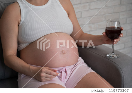 Close-up of the belly of a pregnant woman holding a glass of red wine while sitting on the sofa. Skin rash. Close-up of the belly of a pregnant woman holding a glass of red wine while sitting on the sofa. Skin rash. 119773307