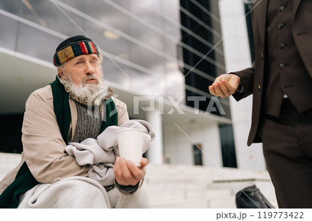Portrait of tired elderly homeless man sitting on urban steps and receiving alms in paper cup from unrecognizable businessman in suit, illustrating social disparities and acts of kindness. 119773422