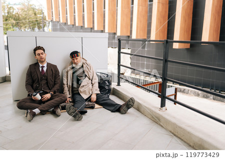 Remote wide shot of chatting young businessman in suit with elderly homeless man sitting on cardboard, representing social and economic disparity. Concept of social issues and poverty. Remote wide shot of chatting young businessman in suit with elderly homeless man sitting on cardboard, representing social and economic disparity. Concept of social issues and poverty. 119773429
