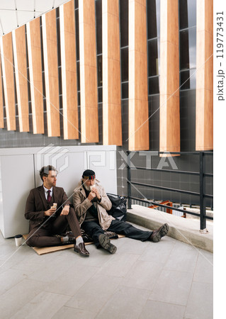 Vertical portrait of kind businessman and elderly homeless man finding common ground as sharing simple meal sitting together on urban street. Concept of social issues and economic disparity. 119773431