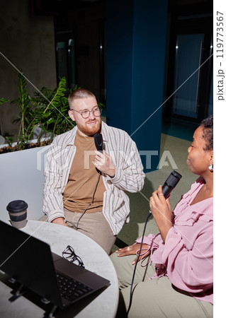 Vertical shot of smiling man in casual outfit co hosting podcast with African American woman while creating content using professional microphones at cafe table, camera flash, copy space 119773567
