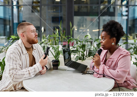 Side view of successful Black woman hosting podcast interviewing businessman while recording broadcast session at cafe table equipped with professional microphones, copy space 119773570