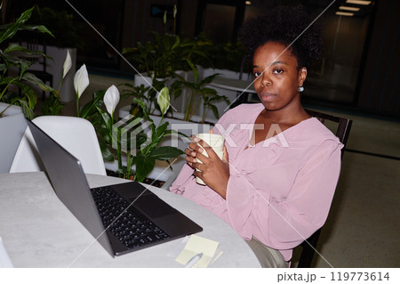 Portrait of serious African American freelance worker during coffee break sitting at table with laptop working remotely from cafe decorated with green plants, camera flash, copy space Portrait of serious African American freelance worker during coffee break sitting at table with laptop working remotely from cafe decorated with green plants, camera flash, copy space 119773614