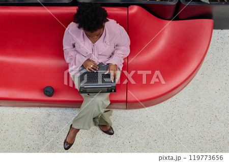 Top view of African American businesswoman typing on laptop keyboard while sitting comfortably on red designer furniture in modern office center, copy space Top view of African American businesswoman typing on laptop keyboard while sitting comfortably on red designer furniture in modern office center, copy space 119773656