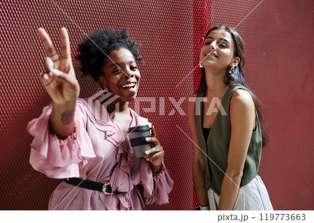Medium shot of two positive female colleagues smiling at camera with V sign posing against red wire grid, while discussing work during coffee break in business center 119773663