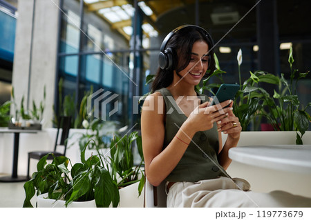 Fashionable woman looking with smile at phone screen enjoying music in headphones while text messaging or writing in social media at cafe table, copy space Fashionable woman looking with smile at phone screen enjoying music in headphones while text messaging or writing in social media at cafe table, copy space 119773679