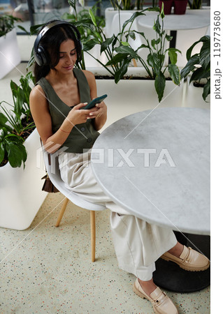 Vertical shot of fashionable woman listening to music in headphones looking at smartphone screen with smile while text messaging or writing in social media sitting at cafe table, copy space Vertical shot of fashionable woman listening to music in headphones looking at smartphone screen with smile while text messaging or writing in social media sitting at cafe table, copy space 119773680