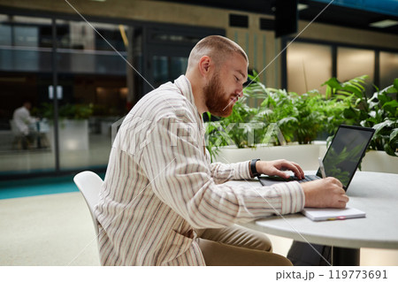 Side view of concentrated adult man using laptop and writing in notebook while studying or working at table in office center cafe, copy space 119773691