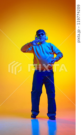 Full-length portrait of stylish elderly bearded man, in sunglasses and cap, listening to music in headphones and dancing against yellow studio background in neon light Full-length portrait of stylish elderly bearded man, in sunglasses and cap, listening to music in headphones and dancing against yellow studio background in neon light 119774209