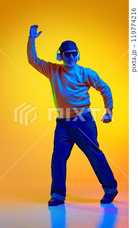 Full-length portrait of stylish elderly bearded man, in sunglasses and cap, listening to music in headphones and dancing against yellow studio background in neon light Full-length portrait of stylish elderly bearded man, in sunglasses and cap, listening to music in headphones and dancing against yellow studio background in neon light 119774216