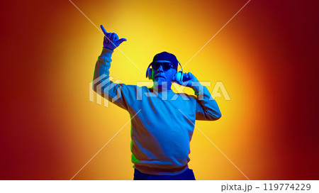 Positive elderly man in sunglasses listening to music in headphones against yellow studio background in neon light Positive elderly man in sunglasses listening to music in headphones against yellow studio background in neon light 119774229