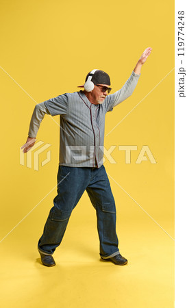 Full-length image of elderly bearded man in cap and sunglasses listening to music in headphones and dancing against yellow studio background 119774248