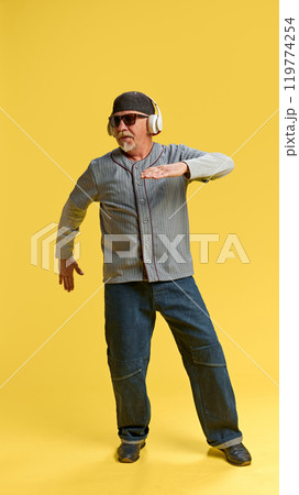 Full-length image of elderly bearded man in cap and sunglasses listening to music in headphones and dancing against yellow studio background 119774254