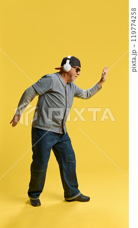 Full-length image of elderly bearded man in cap and sunglasses listening to music in headphones and dancing against yellow studio background 119774258