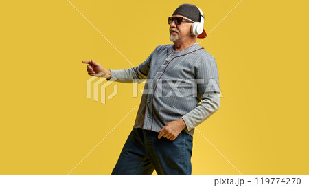 Elderly bearded man in cap and sunglasses listening to music in headphones and dancing against yellow studio background 119774270