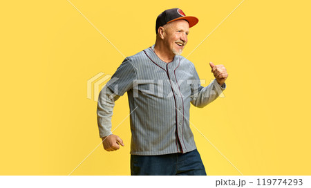 Portrait of happy smiling elderly man in cap cheerfully dancing, showing joy and excitement against yellow studio background 119774293