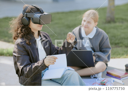 Curly haired girl playing VR game and gesticulating while her blonde bestie watching her Curly haired girl playing VR game and gesticulating while her blonde bestie watching her 119775730