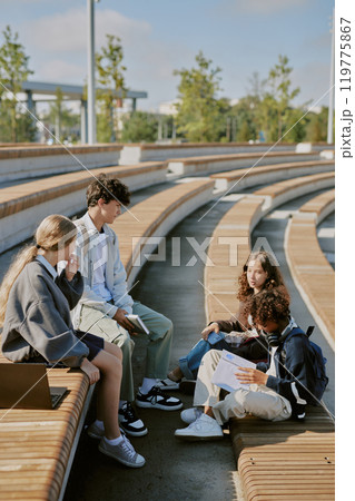 Vertical shot of biracial classmates sitting on wooden bench at stadium and chatting after school day 119775867