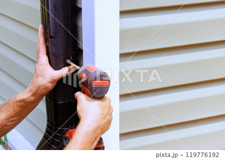 An employee installing rain gutter on roof with is installing downpipe, downspout bend An employee installing rain gutter on roof with is installing downpipe, downspout bend 119776192
