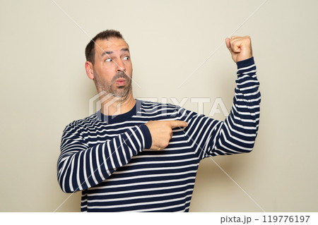 Bearded hispanic man in his 40s wearing a striped sweater showing his pitiful physical condition by pointing at his ridiculous biceps, isolated over beige studio background. Bearded hispanic man in his 40s wearing a striped sweater showing his pitiful physical condition by pointing at his ridiculous biceps, isolated over beige studio background. 119776197