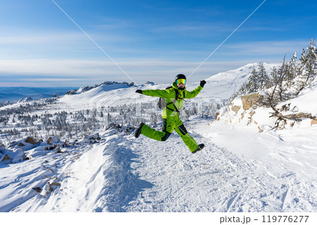 Man jumping Bright acid green outfit: warm suit, goggles, black helmet. snowy mountains background at ski resort. 119776277