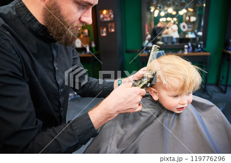 Cute boy excited about new haircut. Blond boy getting stylish haircut in barbershop. Professional hairdresser holding shaver and comb. Cute boy excited about new haircut. Blond boy getting stylish haircut in barbershop. Professional hairdresser holding shaver and comb. 119776296
