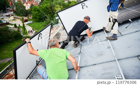 Workers building solar panel system on metal rooftop of house. Three men installers installing photovoltaic solar module outdoors. Alternative, green and renewable energy generation concept. Workers building solar panel system on metal rooftop of house. Three men installers installing photovoltaic solar module outdoors. Alternative, green and renewable energy generation concept. 119776306