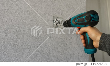 An employee repairing or servicing an electrical outlet or switch using an electric screwdriver against a background of gray wallpaper 119776529
