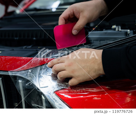 The master applies vinyl film to the headlight of a red car. Closeup view on worker detailer hand smoothing with a scraper protective film.  119776726