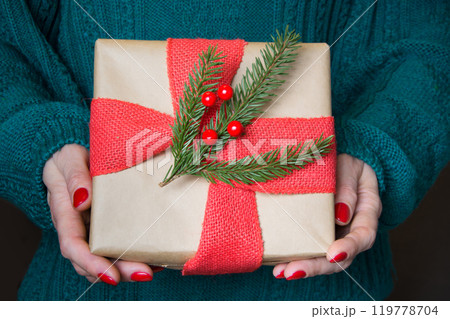 Female holding christmas gift in craft paper with red burlap ribbon with decor. Christmas gifts. Close up. Merry christmas. Boxing Day. Season of giving. Christmas JOY. 119778704