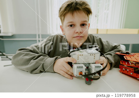 Medium close up portrait of adorable school boy cuddling prototype of robot while sitting at desk 119778958