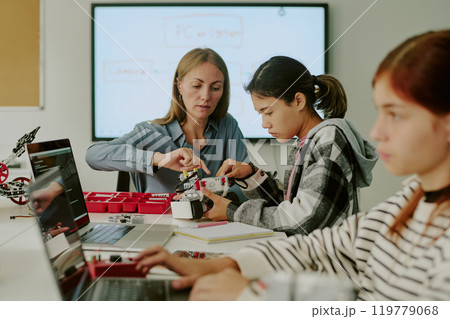 Caucasian teacher and student sitting at work desk and making robot together while other school girl coding 119779068