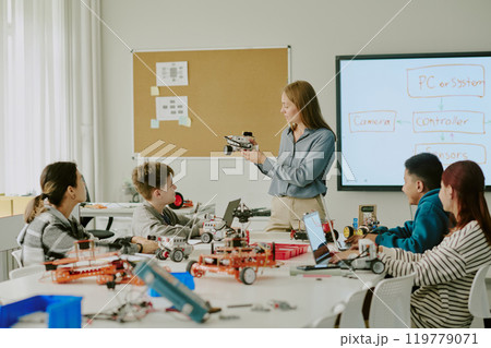 Young teacher showing robot parts while students sitting at desks with robots and listening to teachers explanation 119779071