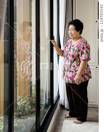 Asian Elderly woman standing by large window, touching glass, looking outside with thoughtful expression, wearing bright patterned shirt and brown pants, barefoot on carpet Asian Elderly woman standing by large window, touching glass, looking outside with thoughtful expression, wearing bright patterned shirt and brown pants, barefoot on carpet 119783543