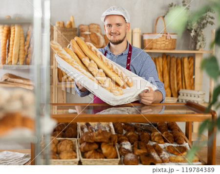 Male seller stands at bakery counter with basket filled by fresh baguettes. Male seller stands at bakery counter with basket filled by fresh baguettes. 119784389