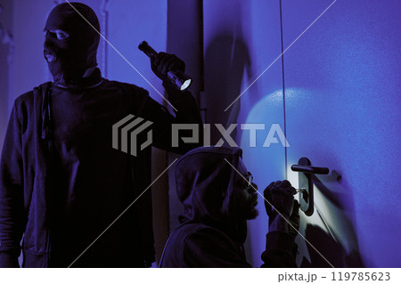Thief in balaclava illuminating door lock and glancing around while his crime partner picking lock Thief in balaclava illuminating door lock and glancing around while his crime partner picking lock 119785623