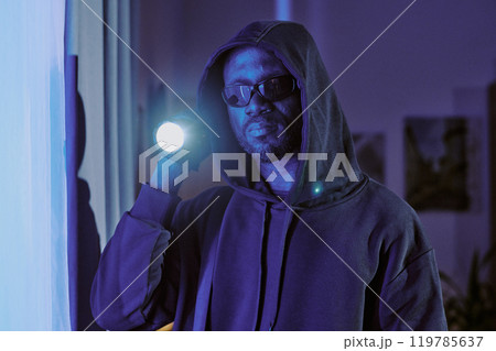 Medium close up portrait of African American thief in black hooded shirt and black glasses illuminating camera with flashlight 119785637